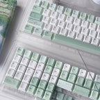 Two mechanical keyboards in clear plastic cases, featuring pastel mint green and white keycaps with delicate floral illustrations, arranged on a white desk alongside a book and a glass object.