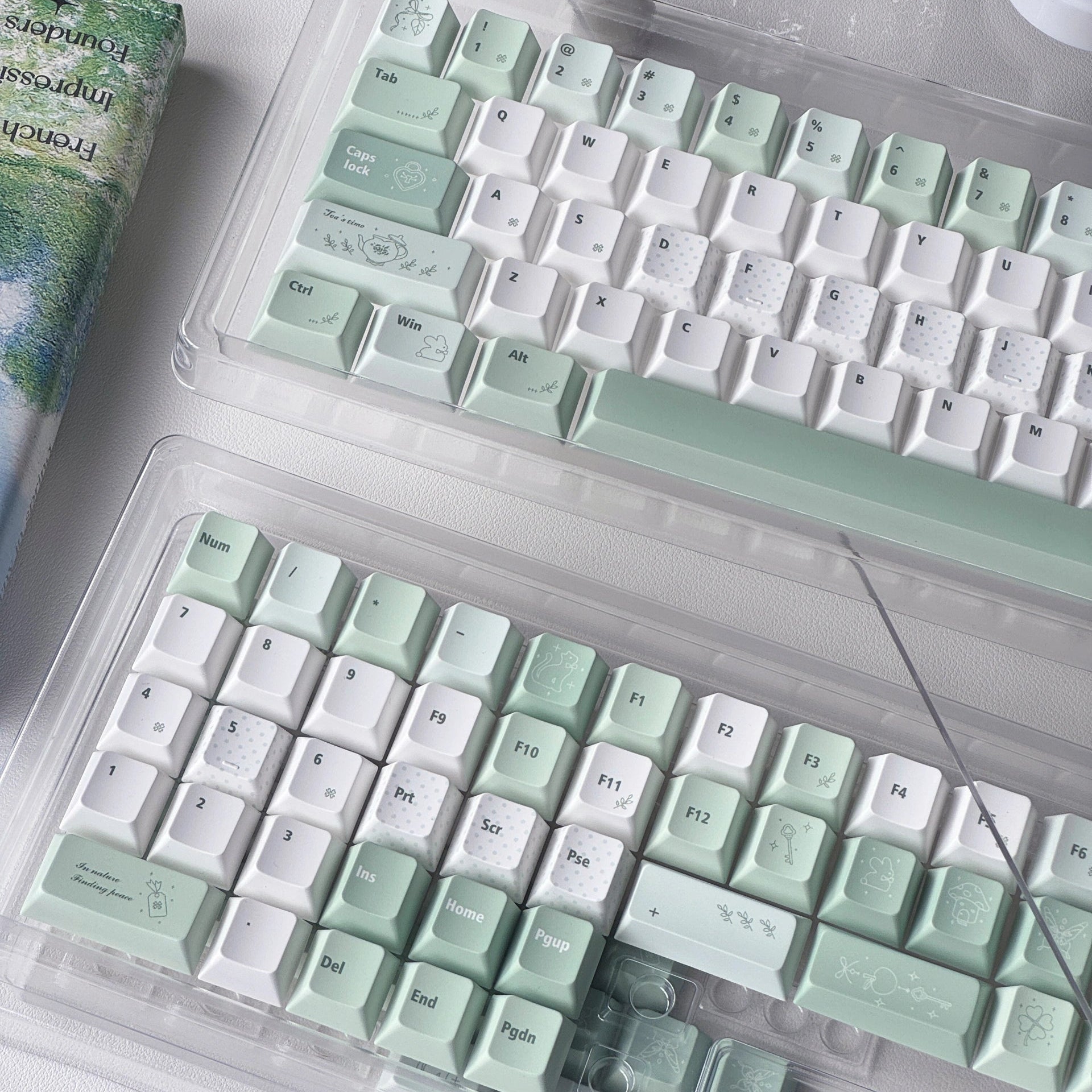 Two mechanical keyboards in clear plastic cases, featuring pastel mint green and white keycaps with delicate floral illustrations, arranged on a white desk alongside a book and a glass object.