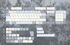A flat-lay view of a keyboard keycap set featuring a pastel, minimalist aesthetic with white, light blue, and gray keycaps arranged over a blurred gray background of trees.