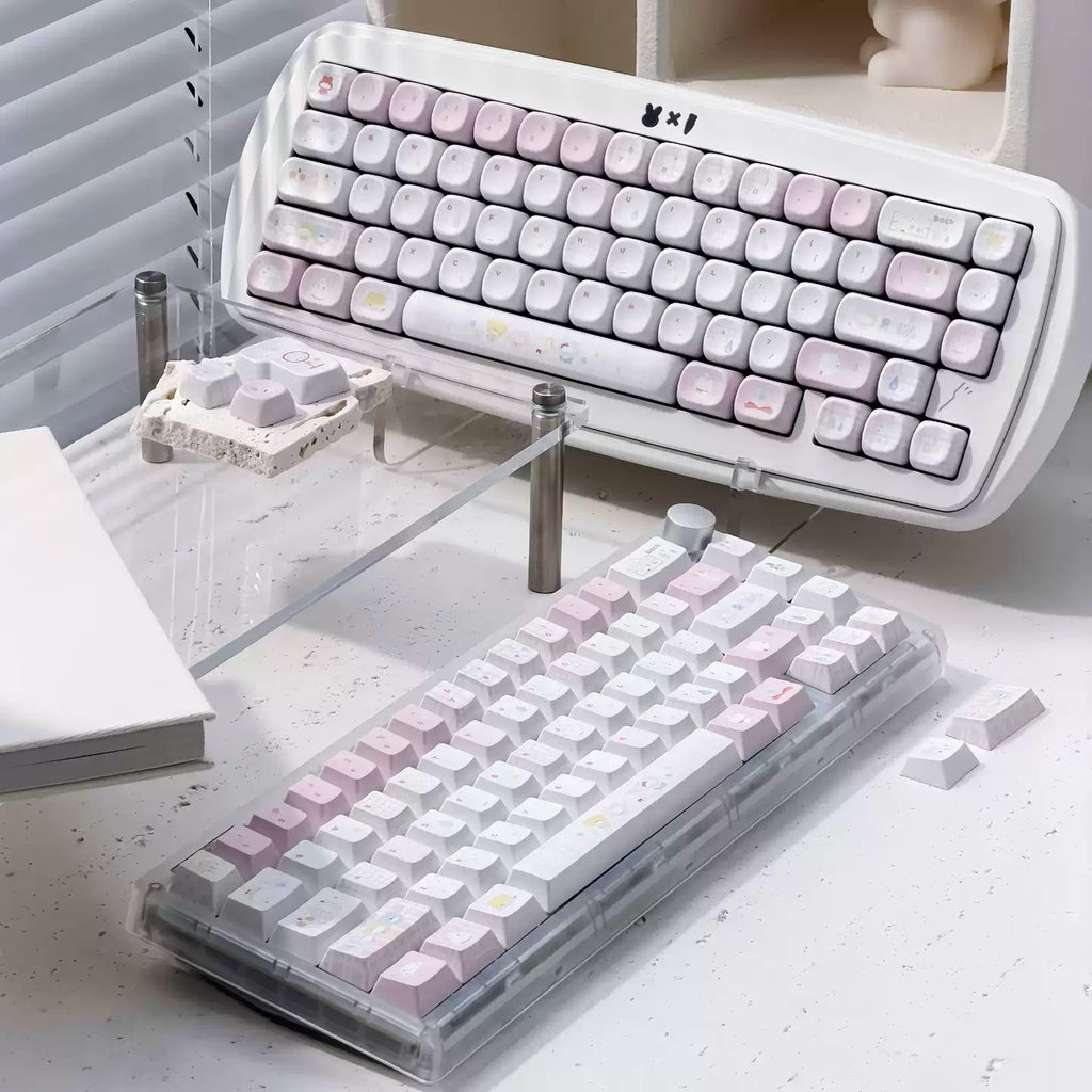 Two mechanical keyboards with pastel pink, white, and light gray keycaps displayed on a desk. One keyboard is shown from a high angle resting on a clear acrylic stand, and the other is shown from a lower angle in a transparent protective case.