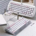 Two mechanical keyboards with pastel pink, white, and light gray keycaps displayed on a desk. One keyboard is shown from a high angle resting on a clear acrylic stand, and the other is shown from a lower angle in a transparent protective case.