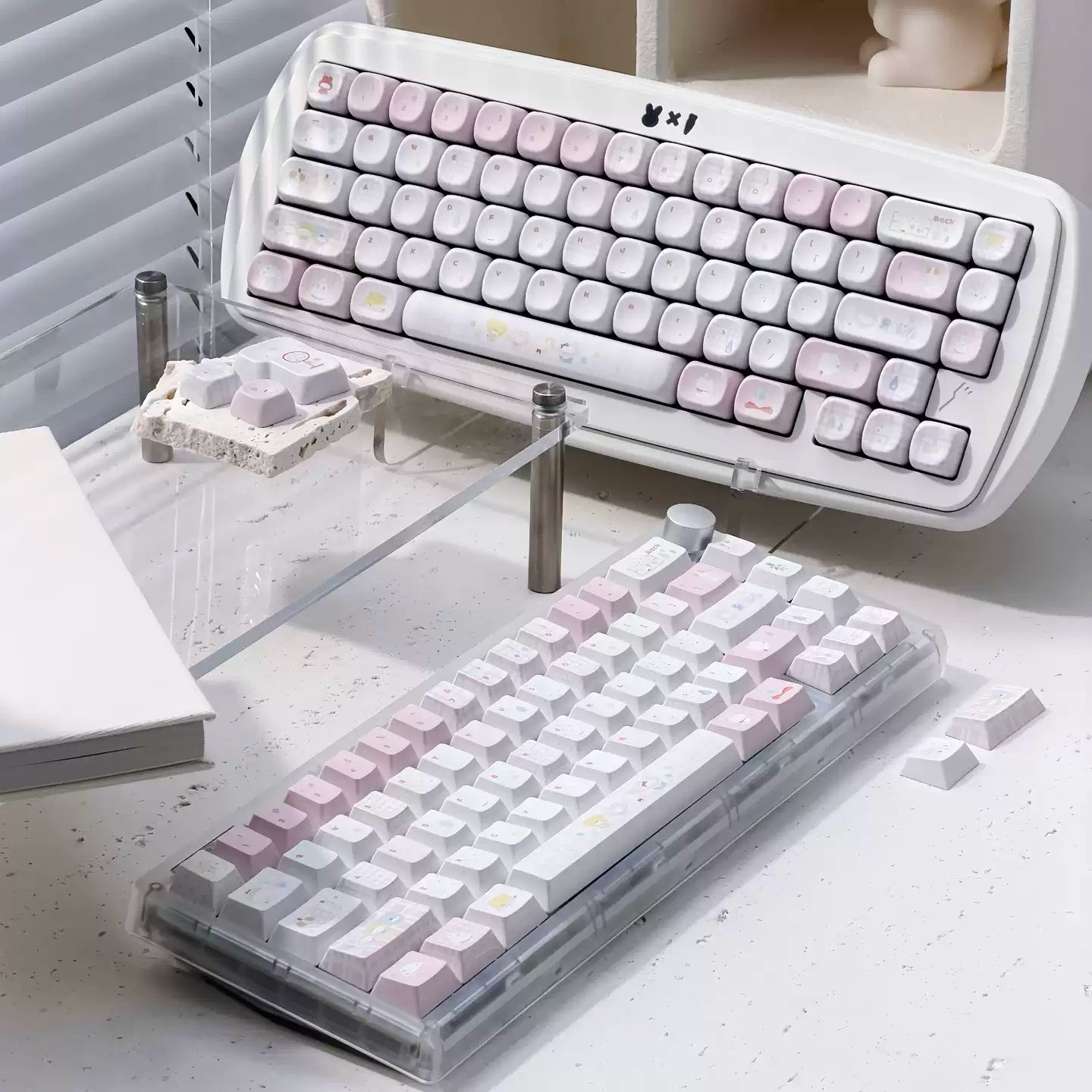 Two mechanical keyboards with pastel pink, white, and light gray keycaps displayed on a desk. One keyboard is shown from a high angle resting on a clear acrylic stand, and the other is shown from a lower angle in a transparent protective case.