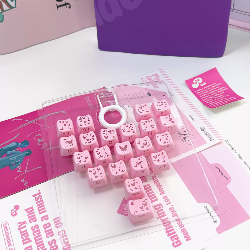 Pink decorative letter keycaps arranged in a clear plastic tray with a white circular key pull, photographed on a white tabletop with pink printed inserts and a purple box in the background.