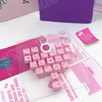 Pink decorative letter keycaps arranged in a clear plastic tray with a white circular key pull, photographed on a white tabletop with pink printed inserts and a purple box in the background.