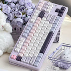 A pastel mechanical keyboard with a lavender body and white, pink, and dark gray keycaps, styled on a desk with purple flowers, a plush toy, and small decorative baskets.