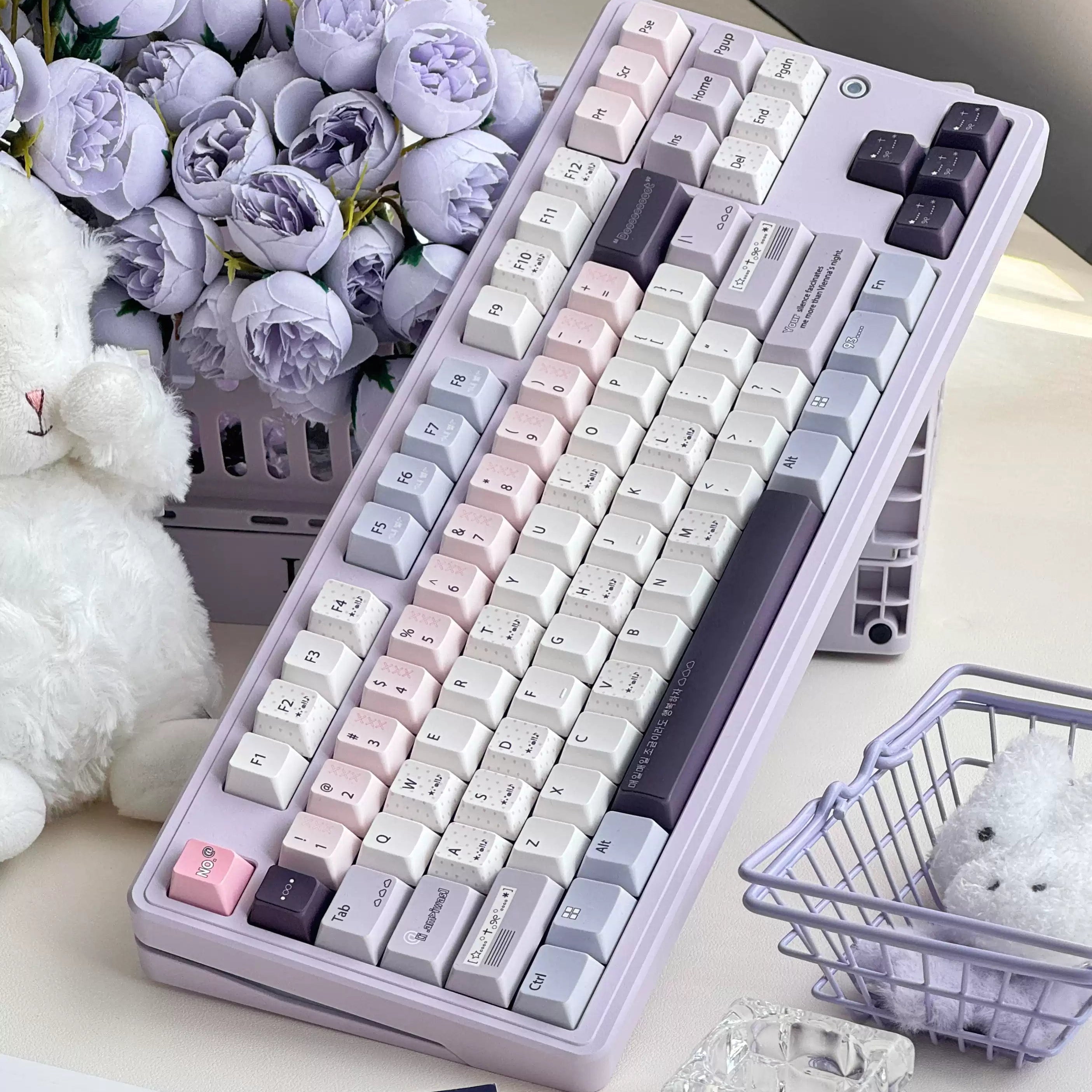 A pastel mechanical keyboard with a lavender body and white, pink, and dark gray keycaps, styled on a desk with purple flowers, a plush toy, and small decorative baskets.