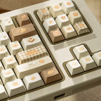 A close-up of a mechanical keyboard with decorative keycaps in warm neutral tones. The keycaps feature pastel cream, beige, and light brown designs, including checkered patterns, small food illustrations, and text such as 'swft'.