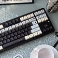 A mechanical keyboard with custom printed keycaps in black, white, and gray, styled on a light blue patterned desk mat next to a white coffee cup and a metal stand holding Tacotac cards.