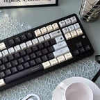 A mechanical keyboard with custom printed keycaps in black, white, and gray, styled on a light blue patterned desk mat next to a white coffee cup and a metal stand holding Tacotac cards.