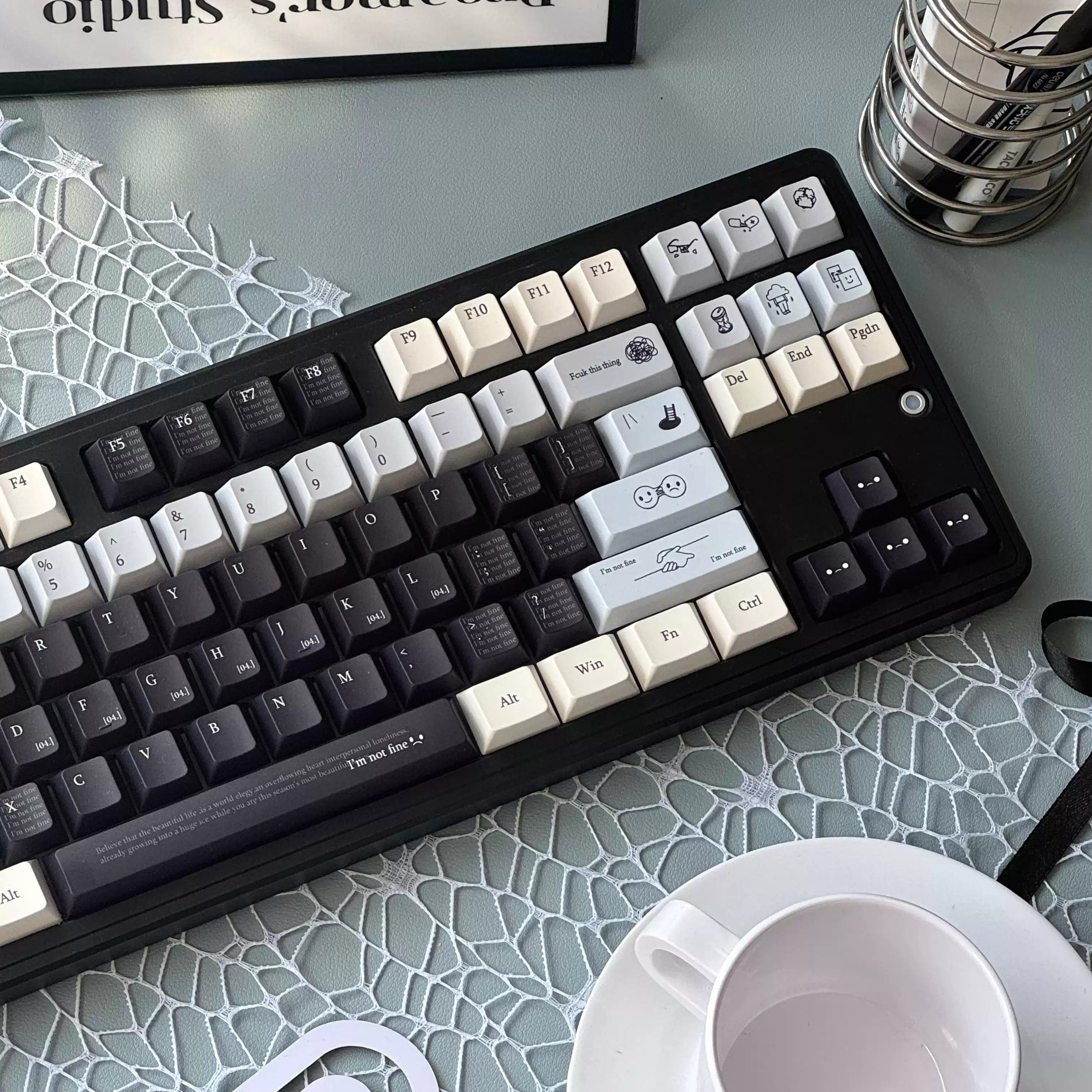A mechanical keyboard with custom printed keycaps in black, white, and gray, styled on a light blue patterned desk mat next to a white coffee cup and a metal stand holding Tacotac cards.