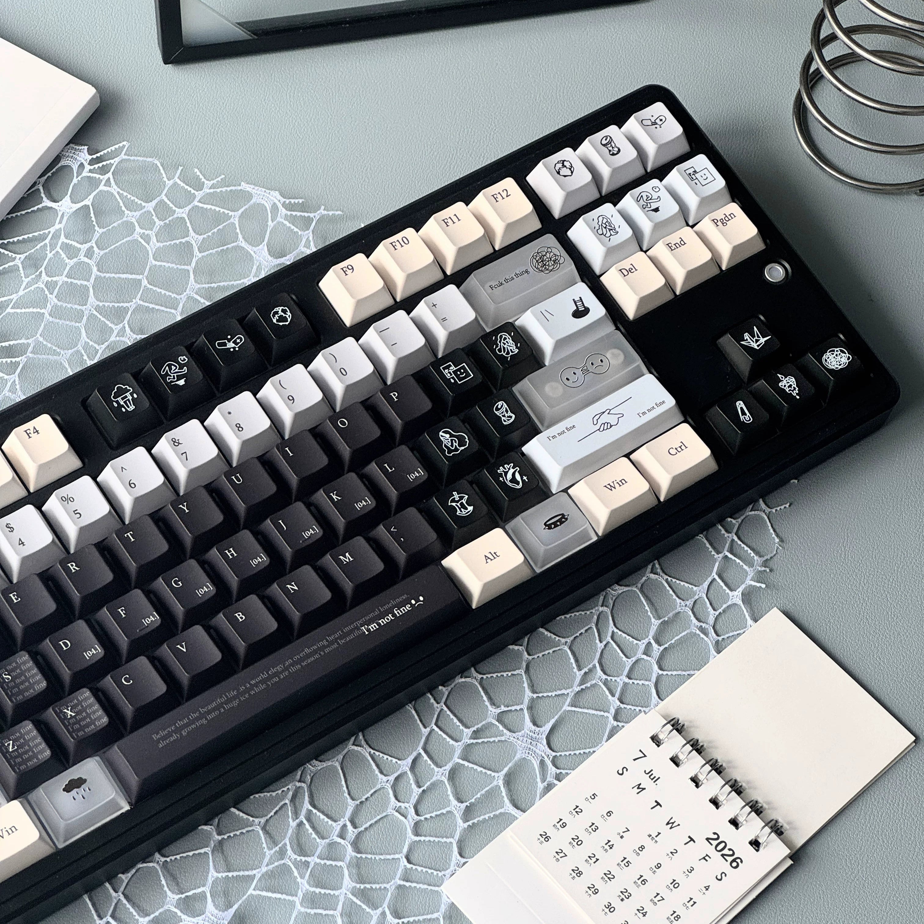 A mechanical keyboard with a black frame and mixed black, white, and beige keycaps, styled on a gray desk with a patterned mat, a small calendar, and other desk accessories.