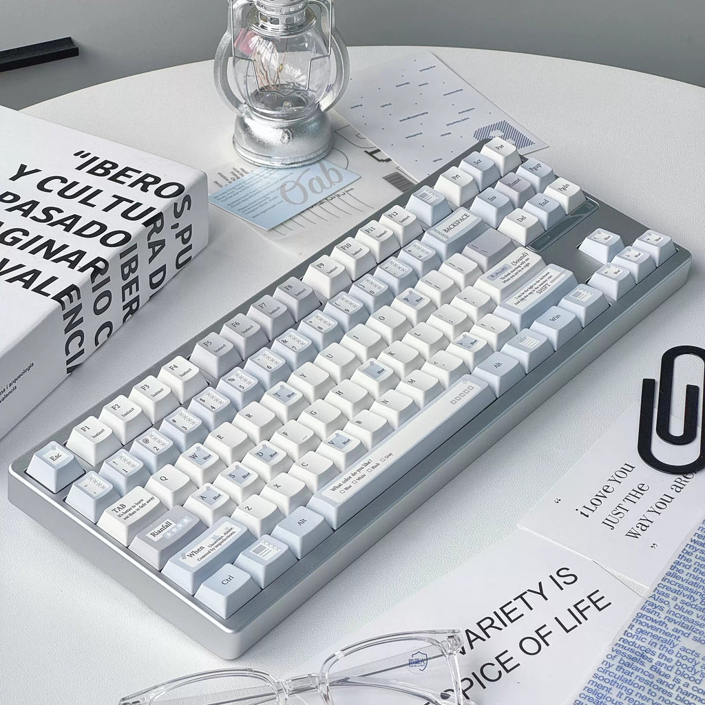 A mechanical keyboard with white and light blue keycaps and a gray frame, styled on a white desk with glasses, a lantern, and printed cards.