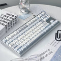 A mechanical keyboard with white and light blue keycaps and a gray frame, styled on a white desk with glasses, a lantern, and printed cards.