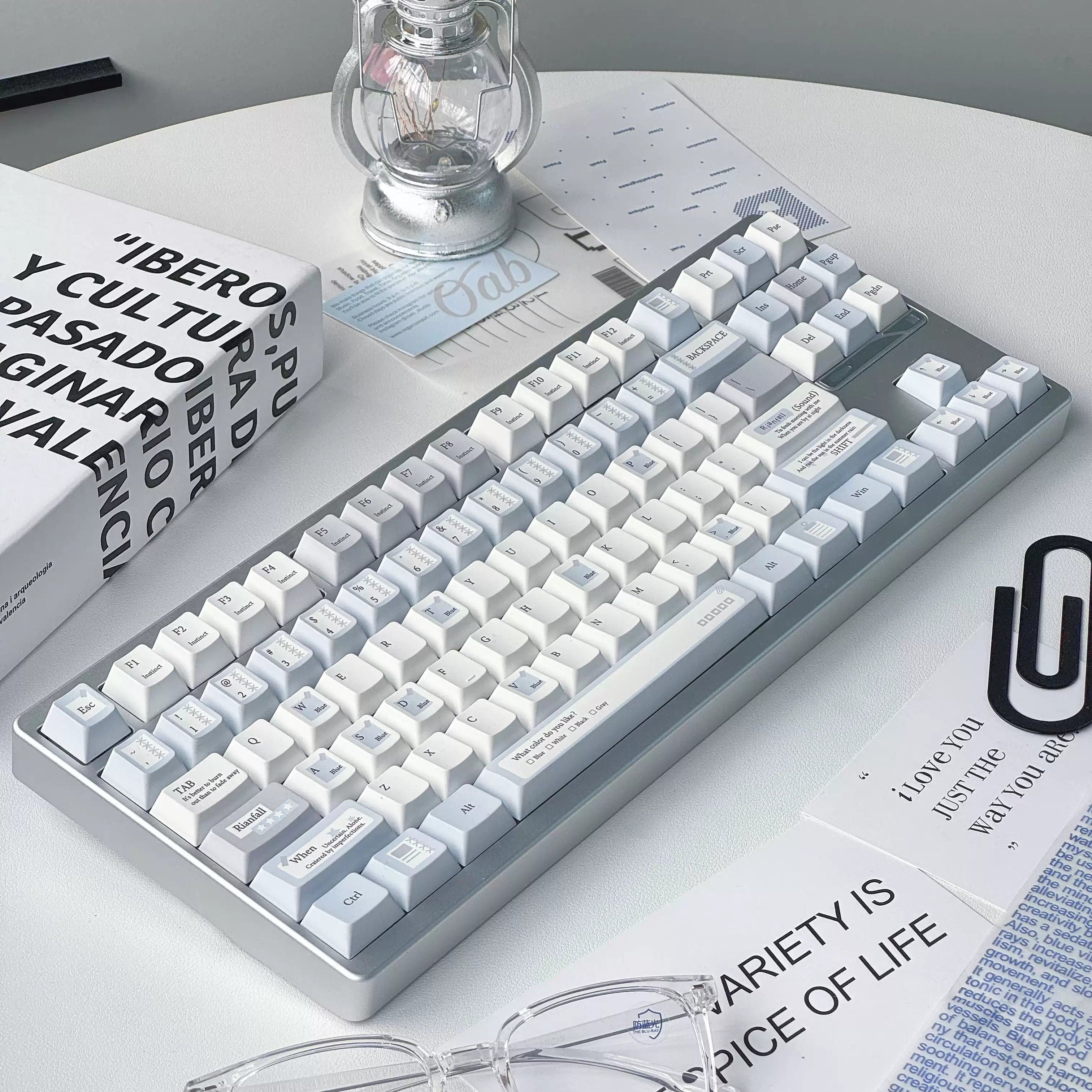 A mechanical keyboard with white and light blue keycaps and a gray frame, styled on a white desk with glasses, a lantern, and printed cards.