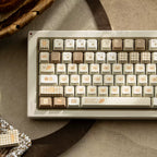 A beige mechanical keyboard with decorative cream, tan, and brown keycaps featuring cute illustrations and checkered patterns, styled on a textured gray surface with dried leaves and a small fabric accessory nearby.