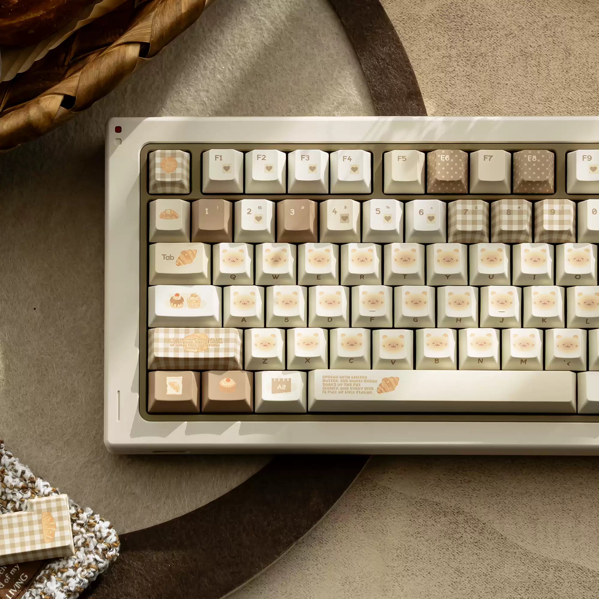 A beige mechanical keyboard with decorative cream, tan, and brown keycaps featuring cute illustrations and checkered patterns, styled on a textured gray surface with dried leaves and a small fabric accessory nearby.