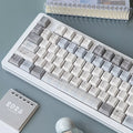 A white mechanical keyboard with pastel gray and cream keycaps, styled on a textured gray desk with notebooks and a 2026 calendar.