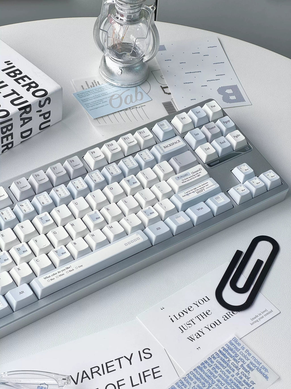 A mechanical keyboard with white keycaps and a light gray frame, styled on a white table with printed cards, a black paperclip, and a glass bulb under a small lamp.