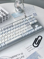 A mechanical keyboard with white keycaps and a light gray frame, styled on a white table with printed cards, a black paperclip, and a glass bulb under a small lamp.