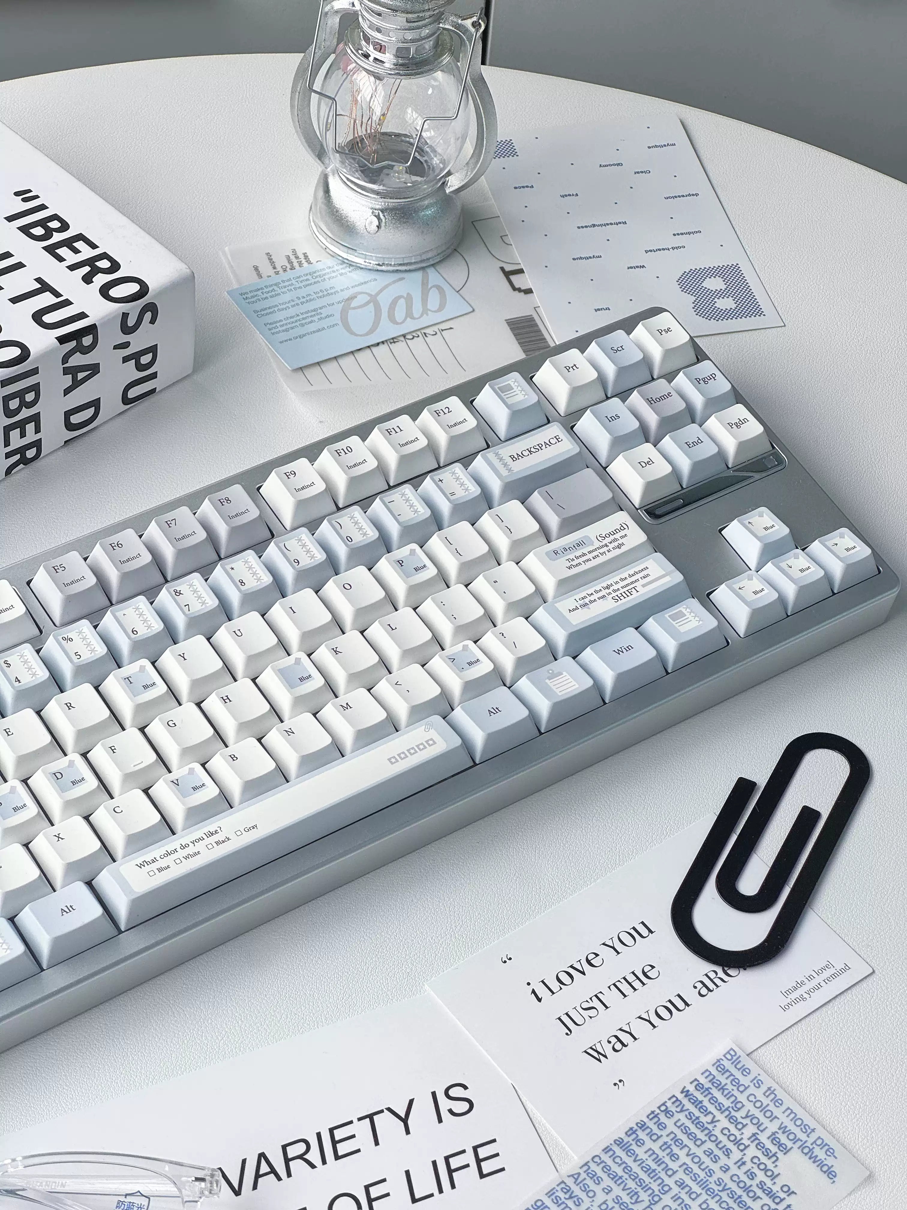 A mechanical keyboard with white keycaps and a light gray frame, styled on a white table with printed cards, a black paperclip, and a glass bulb under a small lamp.