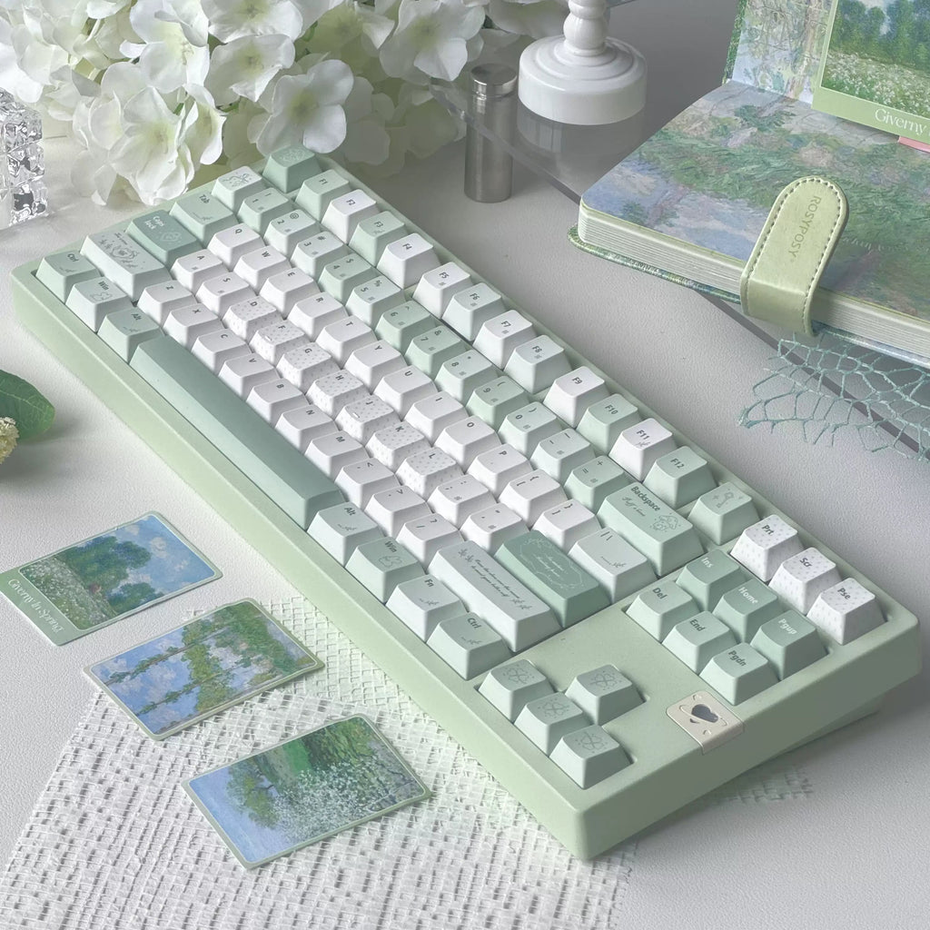 A pastel green and white mechanical keyboard styled on a tabletop with white flowers, a small lantern, a notebook, and decorative cards in a soft, aesthetic setup.