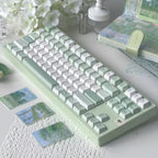 A pastel green and white mechanical keyboard styled on a tabletop with white flowers, a small lantern, a notebook, and decorative cards in a soft, aesthetic setup.