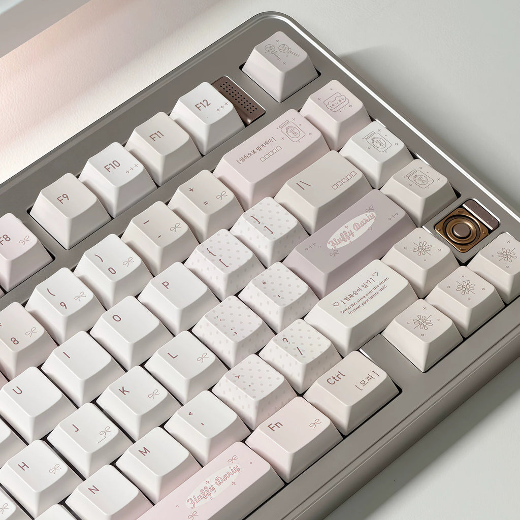 A close-up of a mechanical keyboard with pastel-colored keycaps. The keys transition from white to very light pink and pale lavender, with a metallic silver-gray frame.