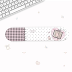 A long, narrow desk accessory with rounded ends, shown on a white desk with a keyboard partially visible in the top-left corner. The product has a white background with black polka dots, a muted mauve plaid section on the left, and a pink heart illustration with a cartoon character on the right.
