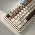 A close-up of a mechanical keyboard with a cream-colored case and keys in a warm brown and beige gradient, featuring custom printed keycaps and a light blue escape key.