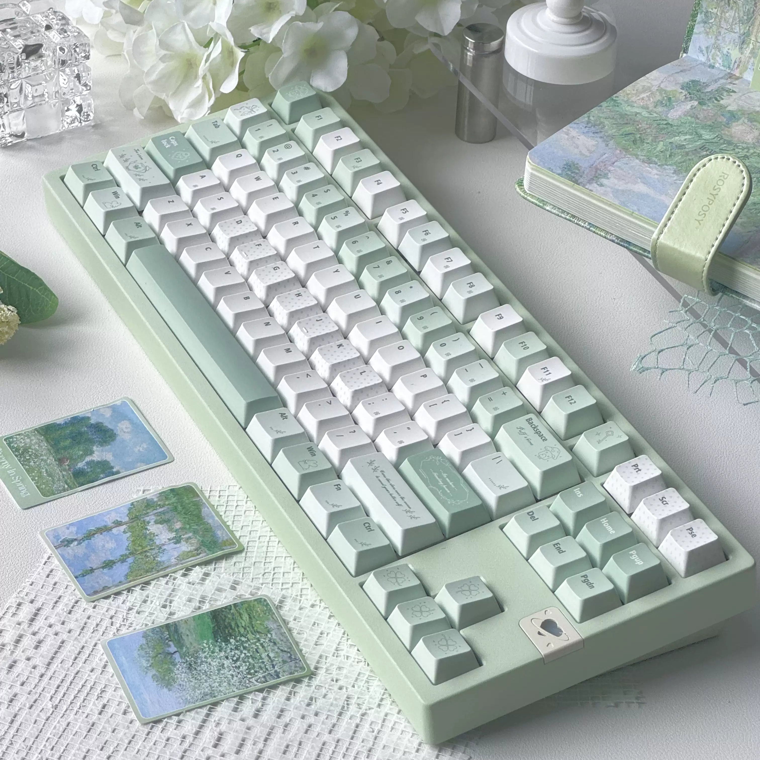 A pastel mechanical keyboard with mint green and white keys arranged on a tabletop beside white flowers, a small lamp, and printed cards with landscape artwork.