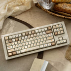 A mechanical keyboard with a cream-colored case and warm beige, brown, and cream keycaps, styled on a textured beige surface with decorative fabric, bread, and a small box.