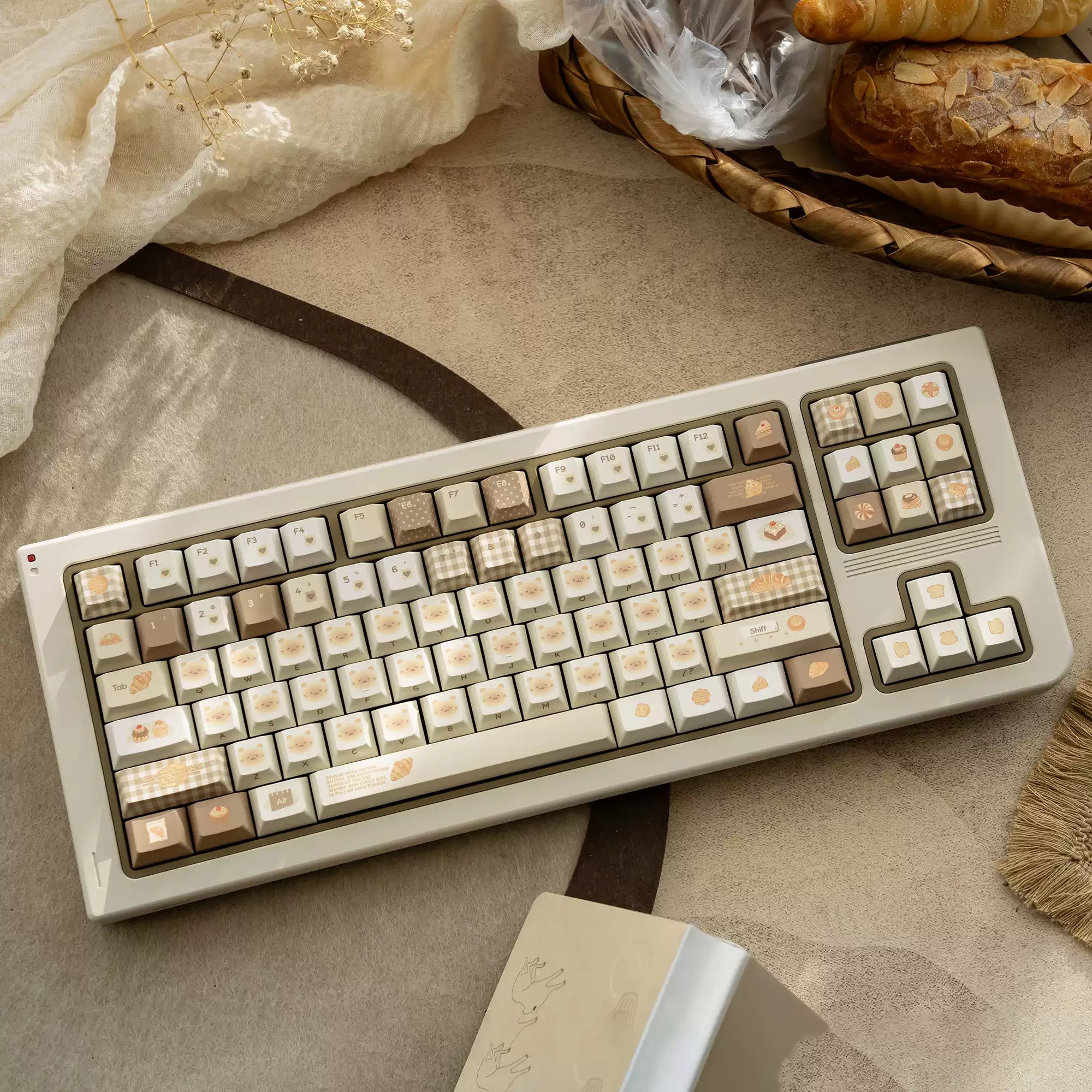 A mechanical keyboard with a cream-colored case and warm beige, brown, and cream keycaps, styled on a textured beige surface with decorative fabric, bread, and a small box.