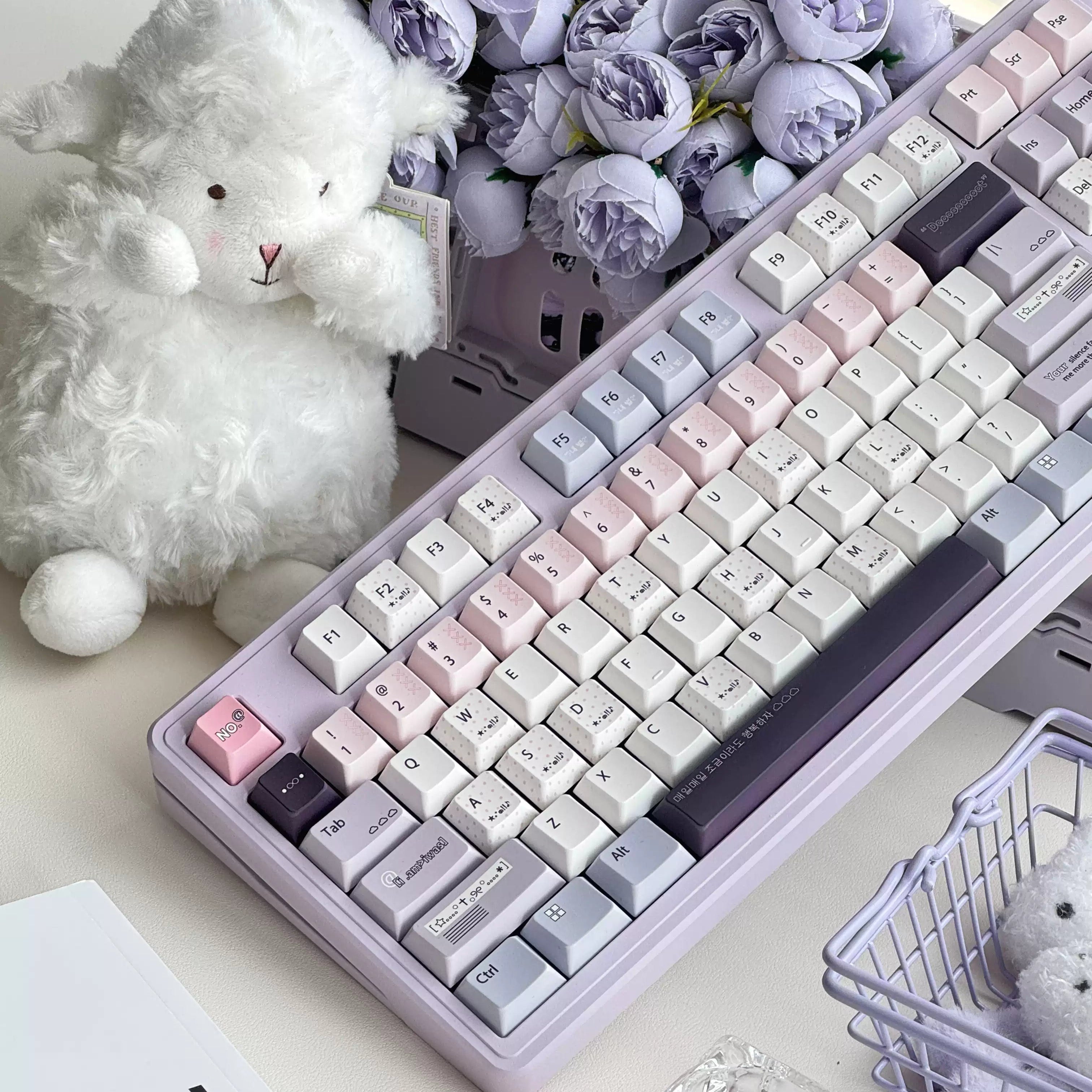 A pastel-colored mechanical keyboard with a lavender chassis and pink, white, and gray keycaps, styled on a desk with a plush white bunny, purple flowers, and decorative props.