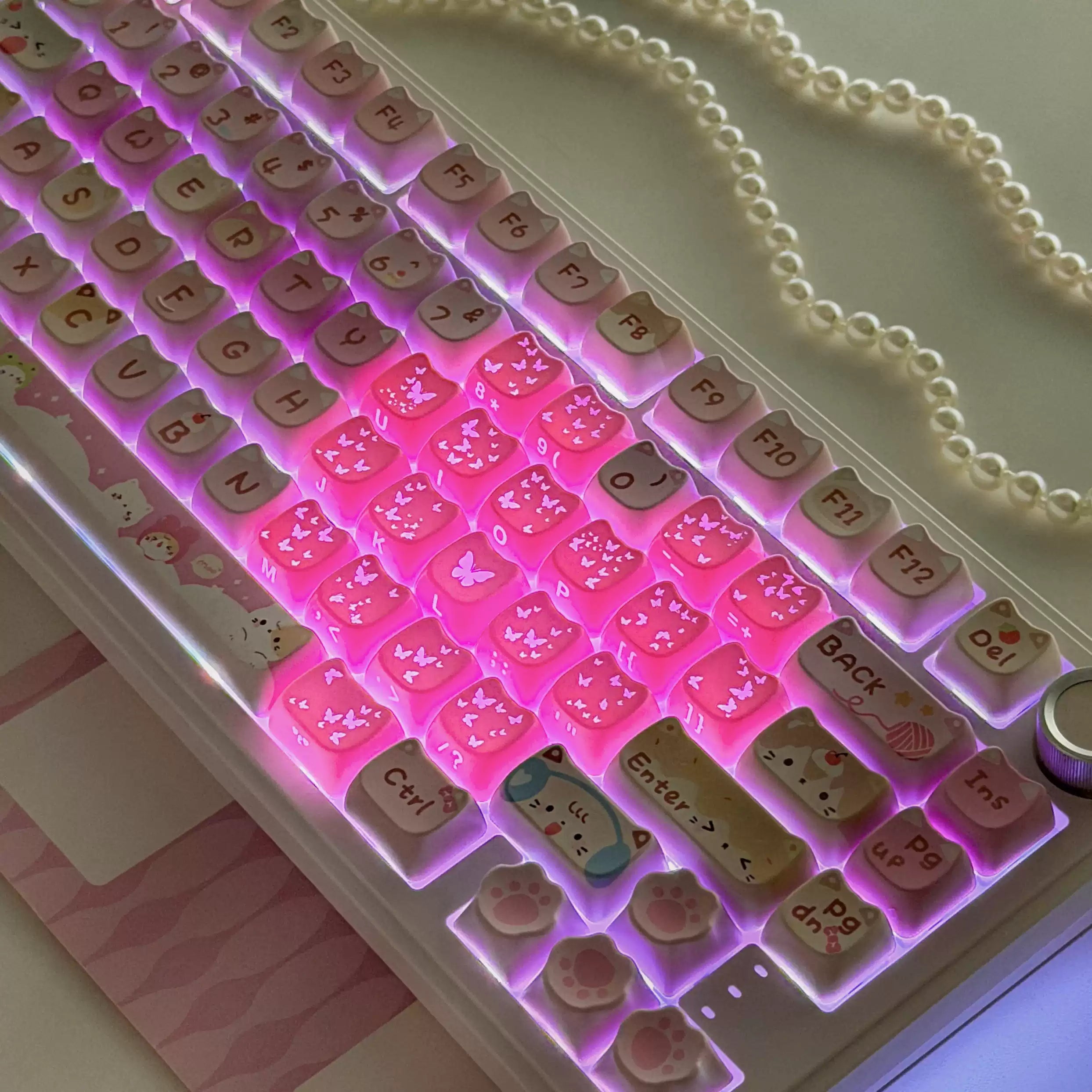 A close-up of a mechanical keyboard with pastel-colored cat-shaped keycaps and pink LED backlighting, shown on a desk with a pearl necklace nearby.