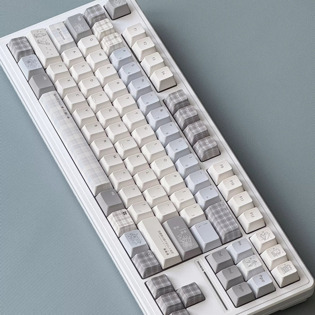 A mechanical keyboard with decorative keycaps in a soft neutral palette of off-white, light gray, and beige plaid patterns, photographed from an overhead angle on a textured slate-blue surface.