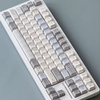 A mechanical keyboard with decorative keycaps in a soft neutral palette of off-white, light gray, and beige plaid patterns, photographed from an overhead angle on a textured slate-blue surface.