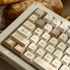 A close-up of a beige mechanical keyboard with decorative food-themed keycaps, including a croissant, cake, and pastry illustrations, with a blurred loaf of bread in the background.