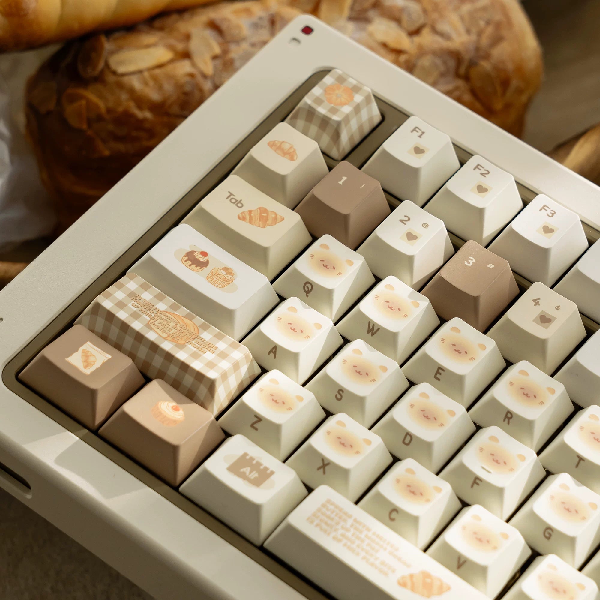 A close-up of a beige mechanical keyboard with decorative food-themed keycaps, including a croissant, cake, and pastry illustrations, with a blurred loaf of bread in the background.