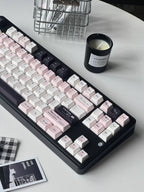 A mechanical keyboard with pastel pink, white, and black keycaps arranged diagonally on a light gray tabletop, alongside a black candle, a metal wire basket, and a small printed card.