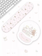 A pastel pink computer mouse pad and matching wrist rest set with cute cartoon angel and teddy bear illustrations, styled on a white desk next to a keyboard.