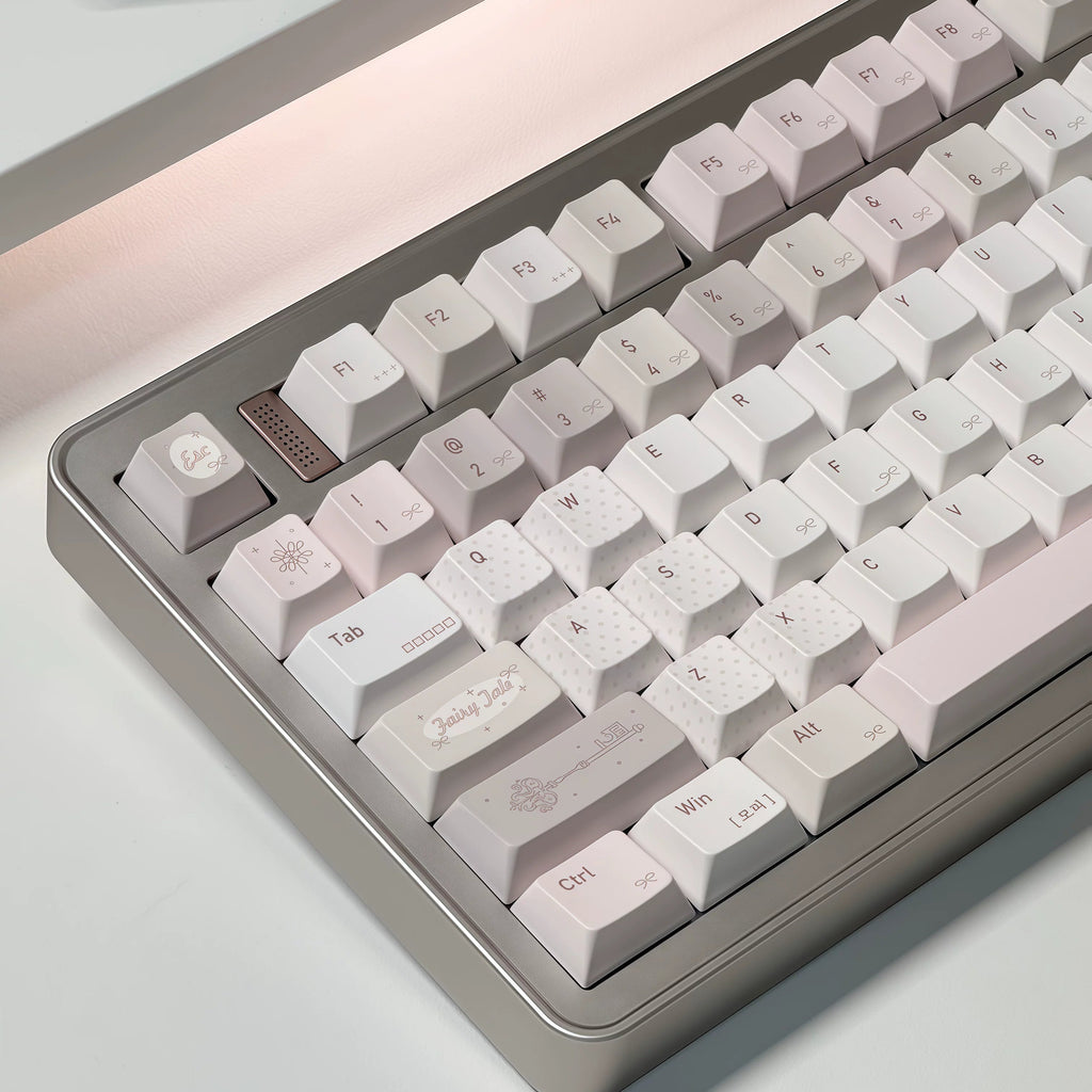 A close-up of a mechanical keyboard with a matte gray frame and pastel white to pale pink keycaps, photographed on a clean light surface with a soft warm light source.