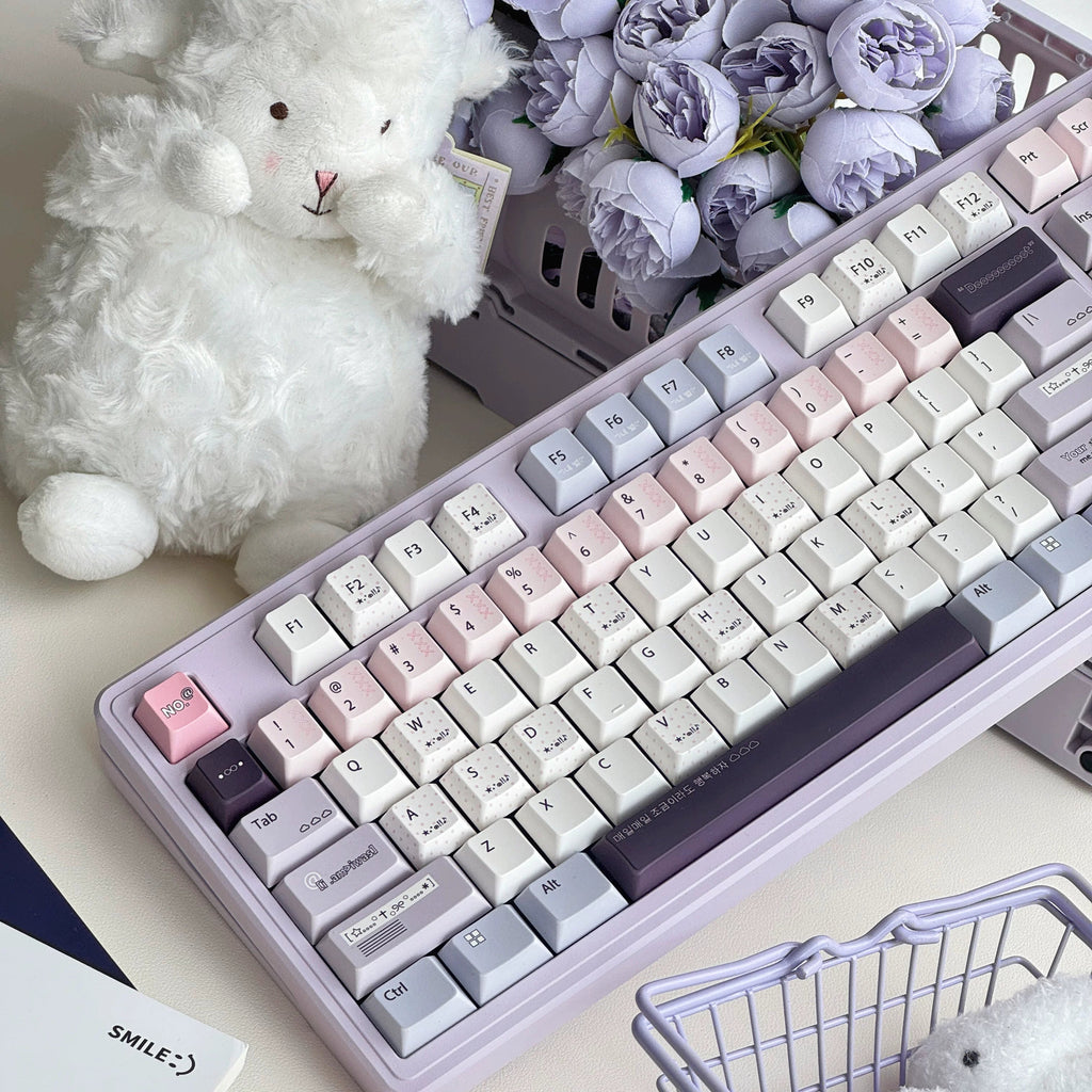 A pastel-colored mechanical keyboard with cream, pink, lavender, and gray keycaps, styled on a desk with a white teddy bear, purple flowers, a small shopping basket, and a clear ice cube.