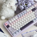 A pastel-colored mechanical keyboard with cream, pink, lavender, and gray keycaps, styled on a desk with a white teddy bear, purple flowers, a small shopping basket, and a clear ice cube.