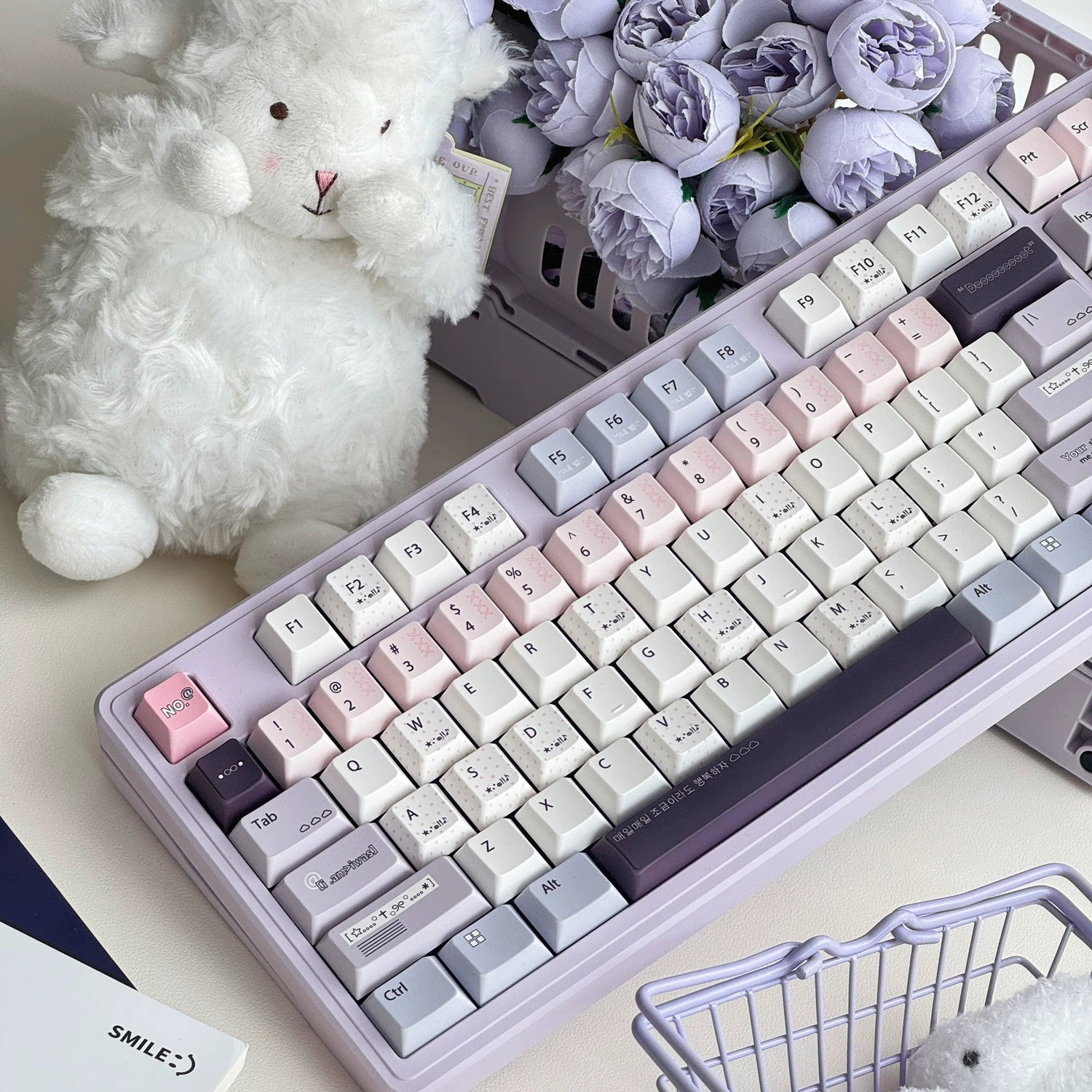 A pastel-colored mechanical keyboard with cream, pink, lavender, and gray keycaps, styled on a desk with a white teddy bear, purple flowers, a small shopping basket, and a clear ice cube.