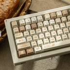 A close-up of a mechanical keyboard with cream, beige, and light brown keycaps decorated with cute pastel illustrations and checkered patterns, styled on a tabletop with a loaf of bread in the background.