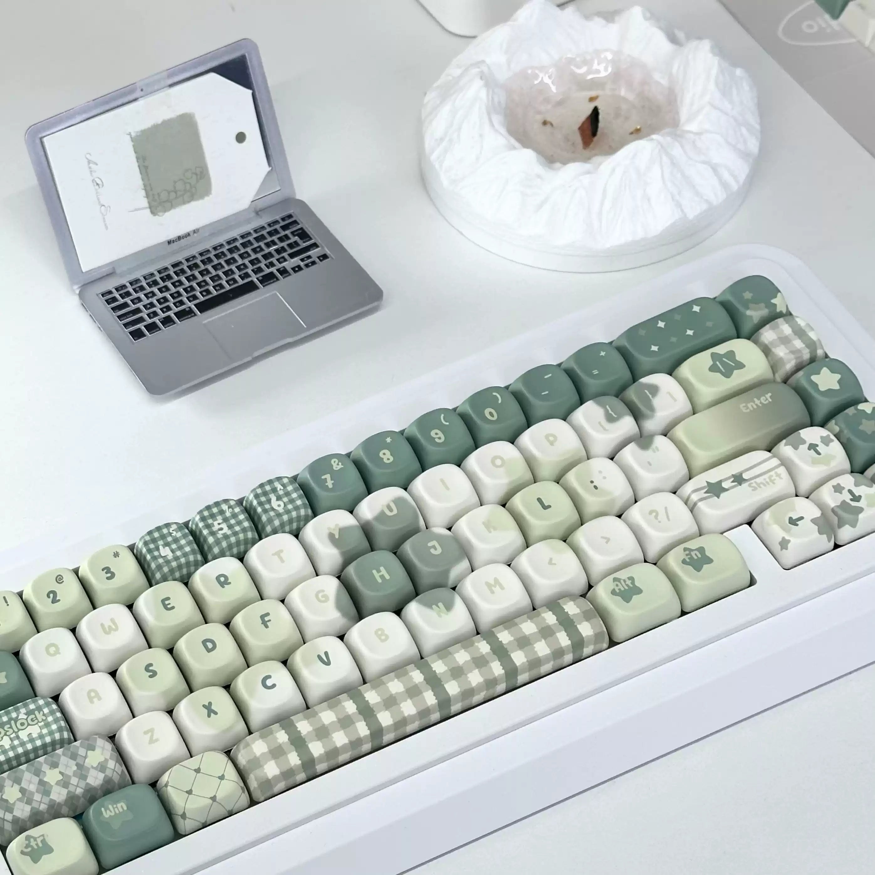 A mechanical keyboard with pastel green, cream, and gray checkered and patterned keycaps, shown on a white desk with a laptop and a white bowl in the background.
