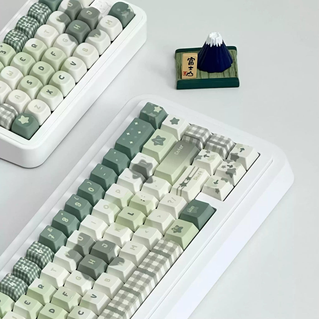A white mechanical keyboard with pastel green, cream, and gray checkered and star-patterned keycaps, shown on a light gray surface. A second keyboard is partially visible in the upper left, and a small dark blue mountain figurine sits near the top center.