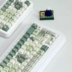 A white mechanical keyboard with pastel green, cream, and gray checkered and star-patterned keycaps, shown on a light gray surface. A second keyboard is partially visible in the upper left, and a small dark blue mountain figurine sits near the top center.