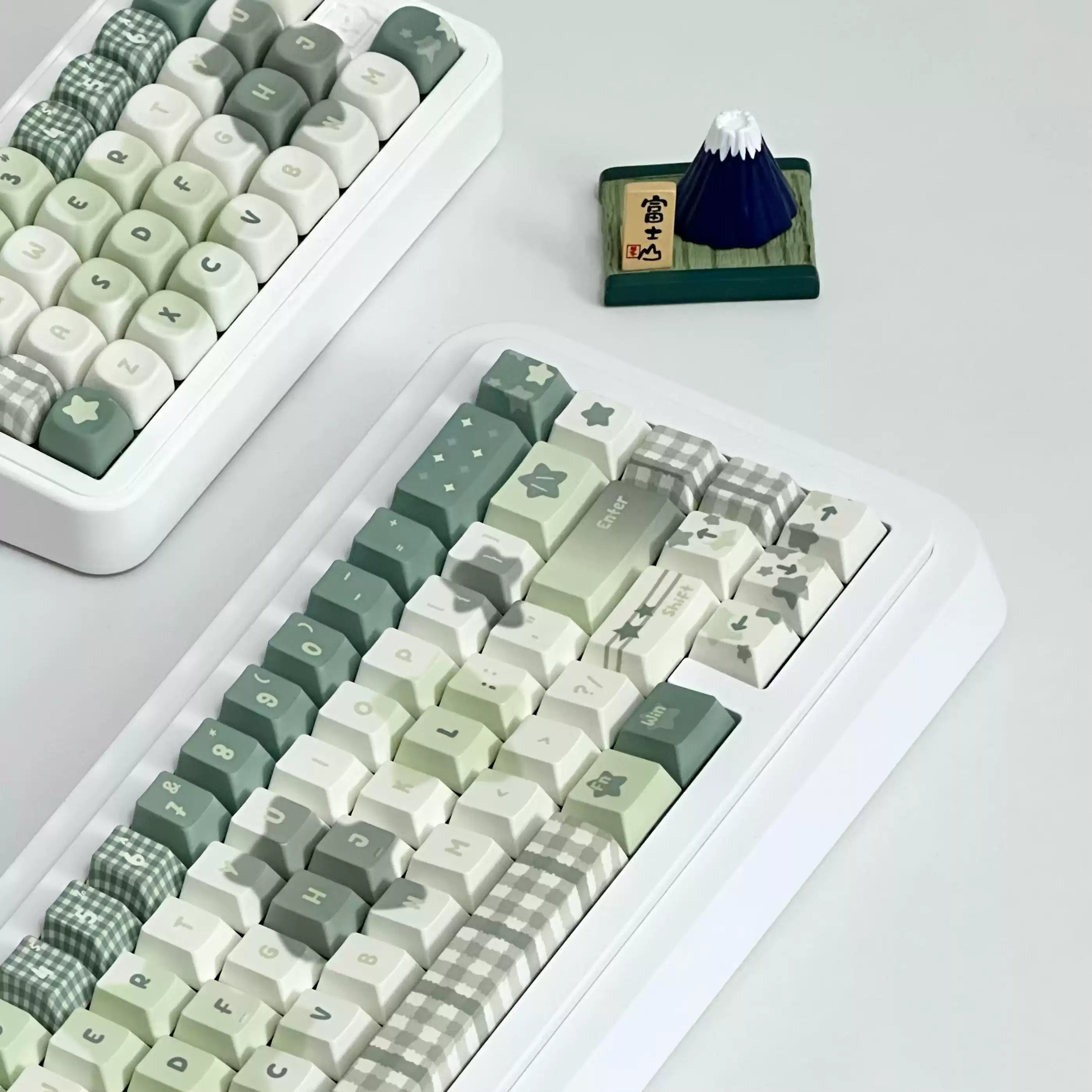 A white mechanical keyboard with pastel green, cream, and gray checkered and star-patterned keycaps, shown on a light gray surface. A second keyboard is partially visible in the upper left, and a small dark blue mountain figurine sits near the top center.