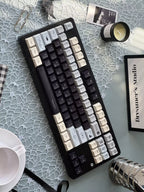 A mechanical keyboard with a black frame and a mix of black, white, and gray keycaps, styled on a light blue patterned surface with desk accessories including a cup, paper clips, and a candle.